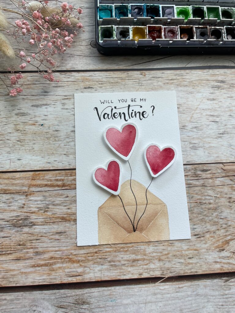 Eine Karte zum Valentinstag mit einem Briefumschlag und dem Spruch "Will you be my Valentine". Aus dem Umschlag ragen mit Aquarell gemalte Herzluftballons heraus, die mit Abstandsklebepads auf dem Aquarellpapier befestigt wurden.