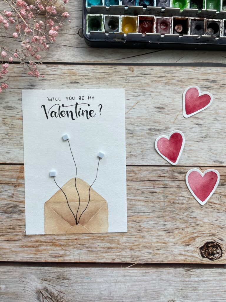 Eine Karte zum Valentinstag mit einem Briefumschlag und dem Spruch "Will you be my Valentine" Es wird gezeigt, wie man die Schnüre für Luftballons mit Fineliner malt.