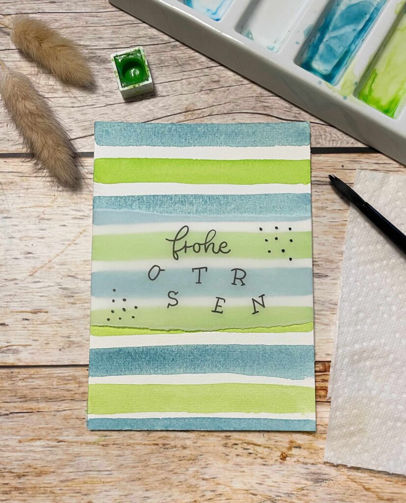 Das Bild zeigt nun die fertige Osterkarte die im Hintergrund mit Aquarell gemalte grüne und blaue Streifen hat und mit dem Spruch "Frohe Ostern" auf Transparentpapier abgerundet wird.