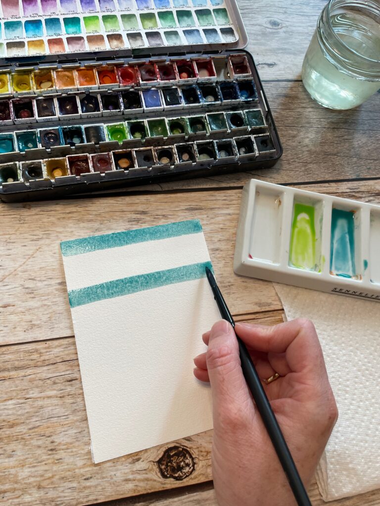 Auf das leere Aquarellpapier nun mit türkiser Aquarellfarbe Streifen gemalt.