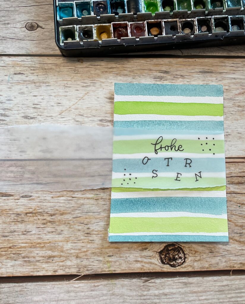 Das mit dem Spruch "Frohe Ostern" beschriftete Transparentpapier wird nun an beiden Seiten um die mit Streifen bemalte Osterkarte herumgeklappt.