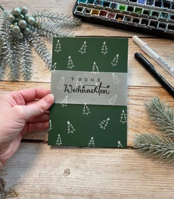 Sandra – Reingemalt – Aquarellmalen lernen, Handlettering und DIY Grußkarten – fünf Inspirationen für einfache DIY Weihnachtskarten (2) Das Bild zeigt eine dunkelgrüne Weihnachtskarte auf der mit einem weißen Gelroller Tannenbäume gezeichnet sind und ein Weihnachtsgruß mit Transparentpapier oben drauf geklebt ist.