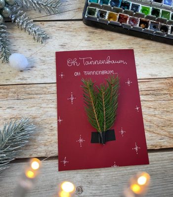 Sandra – Reingemalt – Aquarellmalen lernen, Handlettering und DIY Grußkarten – fünf Inspirationen für einfache DIY Weihnachtskarten 3# Das Bild zeigt eine dunkelrote Weihnachtskarte mit zwei Tannenzweigen darauf.